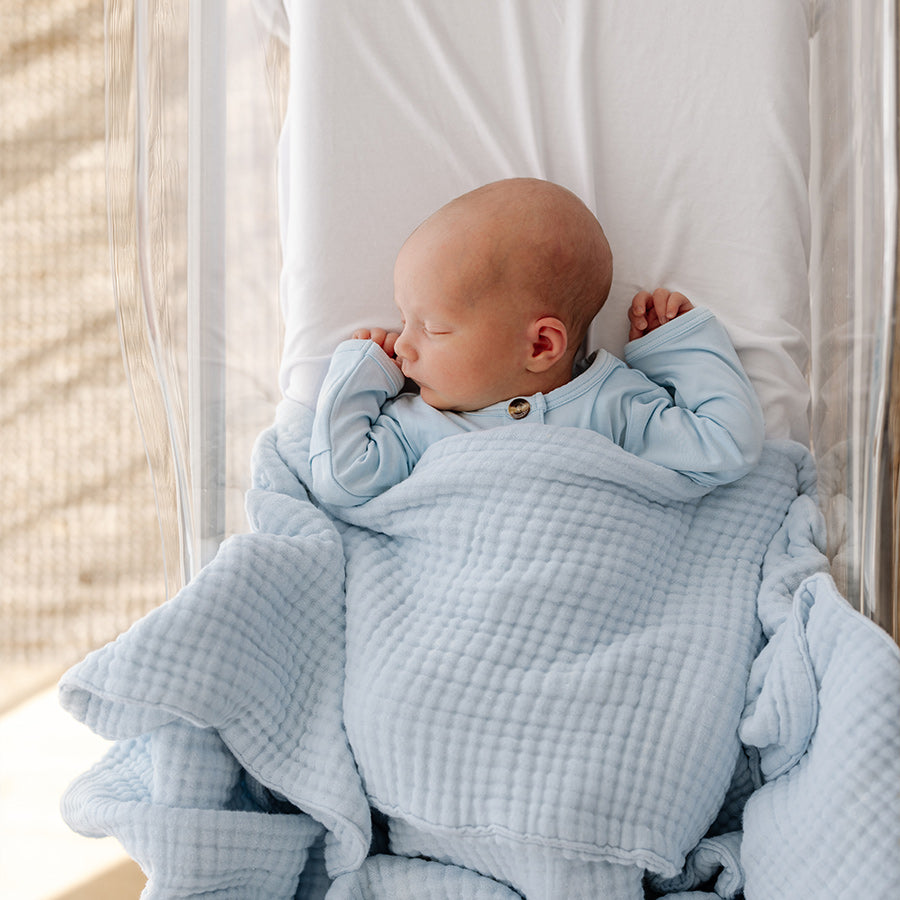 baby sleeping in clear basinet, comfortably wrapped in a bubble textured muslin blanket and matching blue gown. 