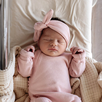 Baby comfortable in basinet lined with cream sheet and cotton blanket wearing a matching pink headband and one-piece-suit.