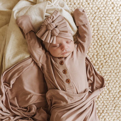 Baby stretching while laying on thick carpet, wrapped in brown swaddle wearing a 3 knot turban. 