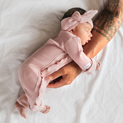 Baby girl leans on father's tattooed arm wearing ribbed pink headband and onesie. 