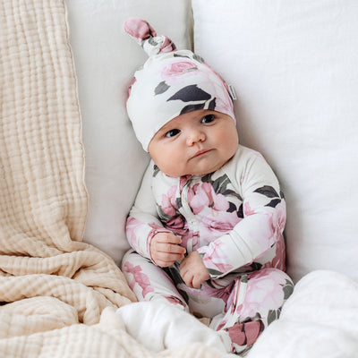 Baby in floral onesie and matching top knot beanie, sitting on a cream blanket, looking ahead.