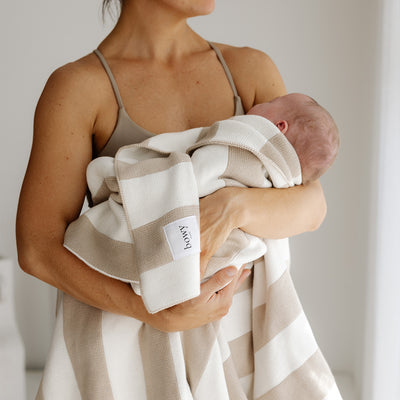 "A mother holding her newborn baby wrapped in a soft, neutral-toned knit blanket with thick white and beige stripes. The blanket features a visible Bowy Made label. The scene is set in a bright, minimalistic room with soft natural lighting.