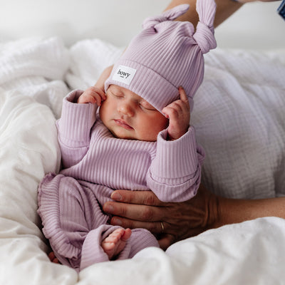 Adorable baby being placed on white blanket wearing ribbed cotton set with delicate frill in lilac. 