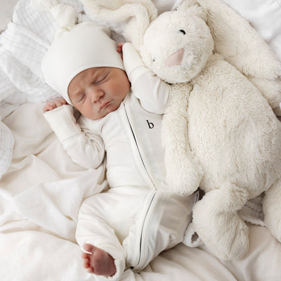 Sleeping baby in white vanilla onesie and beanie, cuddling a plush bunny, resting on soft white bedding.