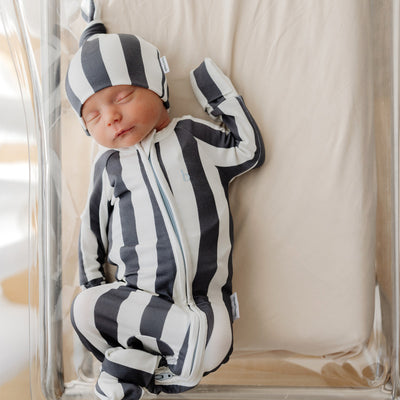 A newborn sleeps peacefully in a clear bassinet, wearing a SoftLuv onesie with navy blue and white stripes and a matching knotted beanie. The outfit features a double zip and fold-over cuffs, resting on neutral-toned bedding.