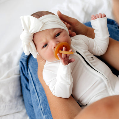 Newborn in white onesie with zipper and bow headband, using a rubber pacifier.