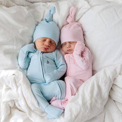 Two newborn babies looking very comfortable wearing matching onesies and top knot beanies, one in pink and another in blue.  