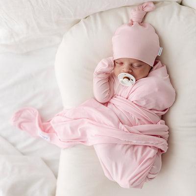 Baby sleeping on white pillow in well lit room, wrapped in light pink swaddle wearing a top knot beanie of the same colour. 