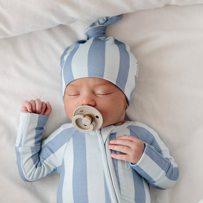 Newborn sleeping in a blue striped onesie with a matching knotted beanie and pacifier.