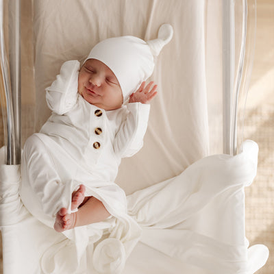 happy baby wearing a white knotted gown with a top knot beanie of the same colour, in a well lit room. 