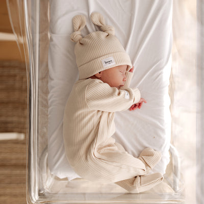 baby asleep in clear cot wearing cream ribbed onesie and wearing matching cream ribbed double knot beanie 