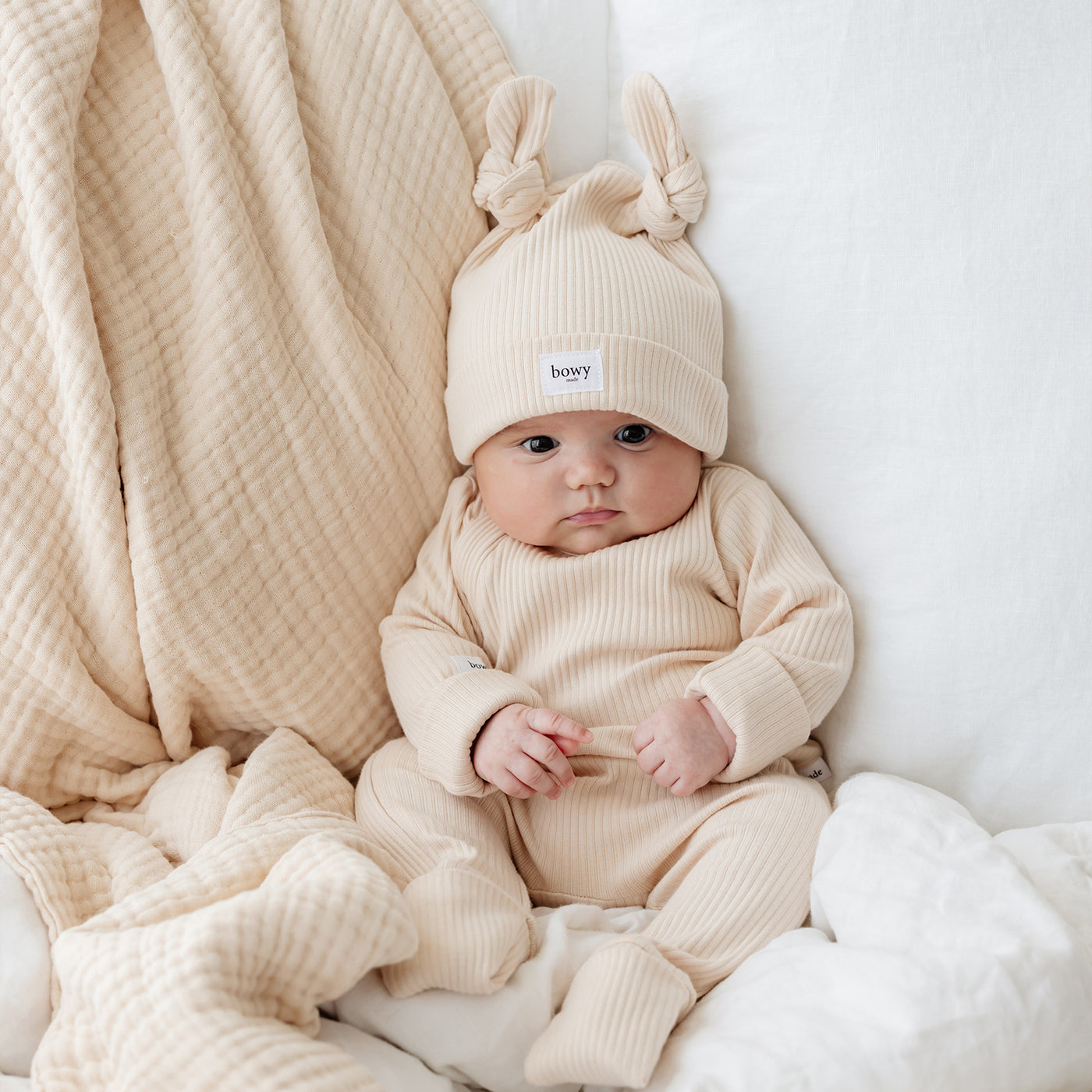 baby boy sitting upright amongst blankets wearing cream onesie set with matching double knot beanie.