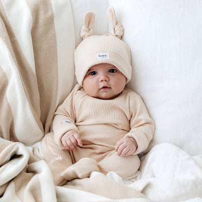 3 month old baby girl wearing cream beanie with onesie - amongst white pillow and soft strip cotton blanket. 