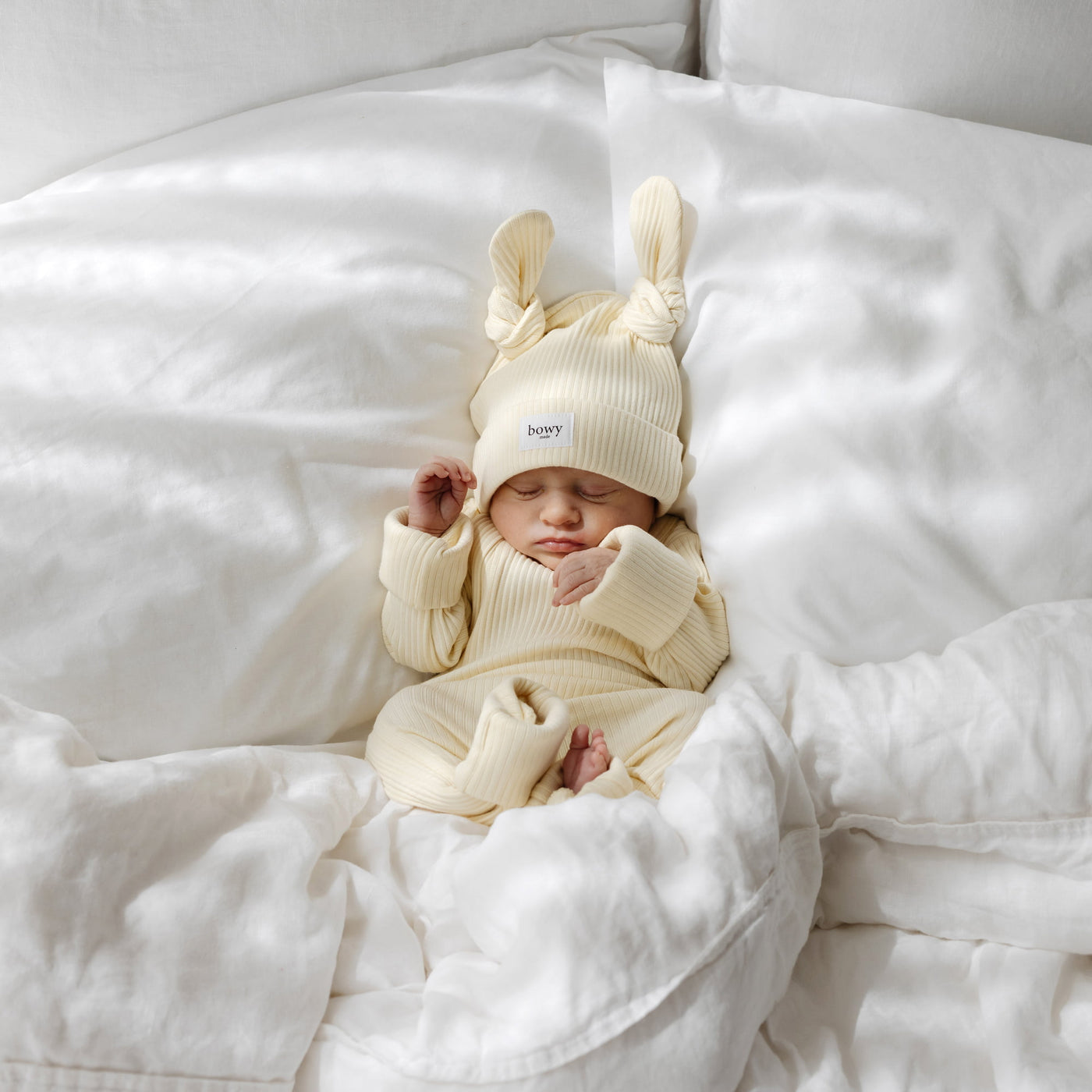 Newborn baby in a yellow-coloured outfit with double knot beanie on a white blanket
