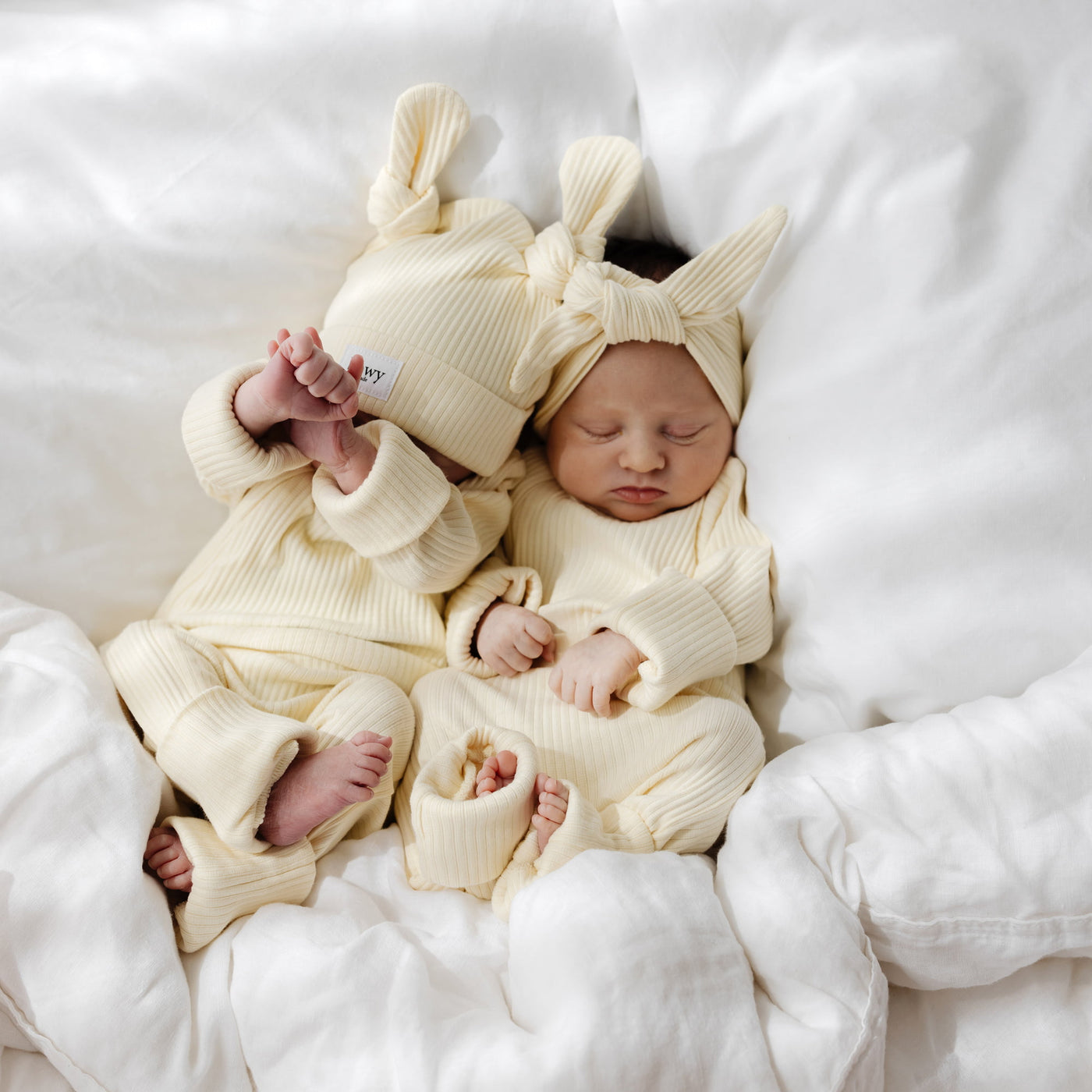 Two newborn babies in matching outfits and with double knot beanies lying on a white blanket.