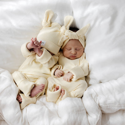 Two newborn babies in matching outfits and with double knot beanies lying on a white blanket.