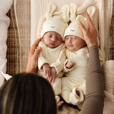 Two babies in matching outfits with matching top knot beanies, held by a mother.