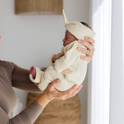 Newborn baby wearing matching yellow headband and onesie being held by a person in a neutral setting.
