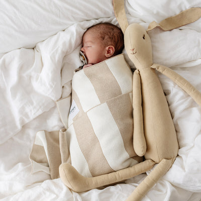 Baby wrapped in striped blanket sleeps on white linen alongside long bunny doll