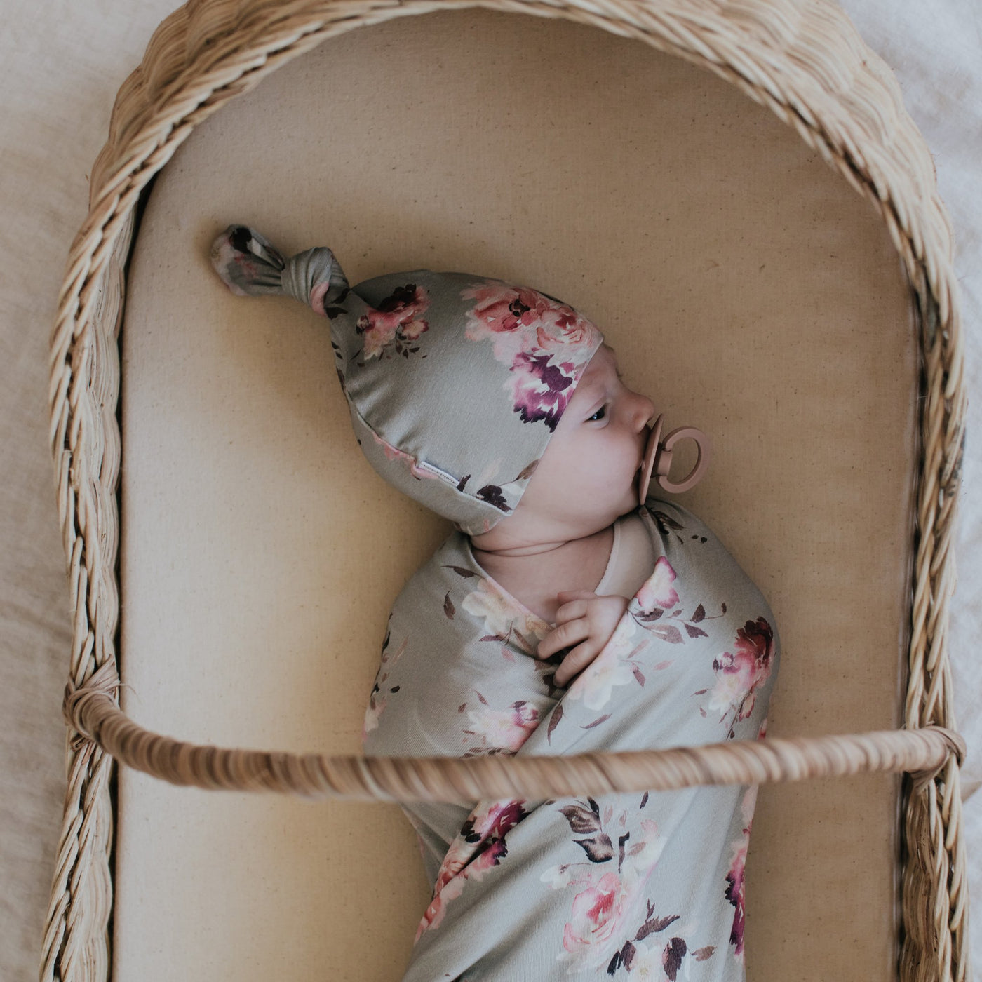 Baby laying in bassinet wearing matching floral swaddle and top knot beanie