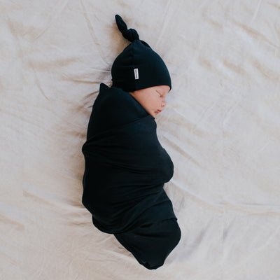 newborn baby in matching navy swaddle and top knot beanie lying on linen sheet